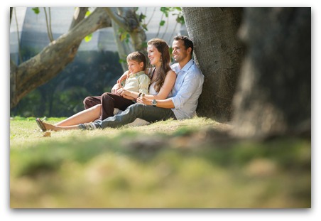 Happy family with man, woman and child leaning on tree in city park. Copy space